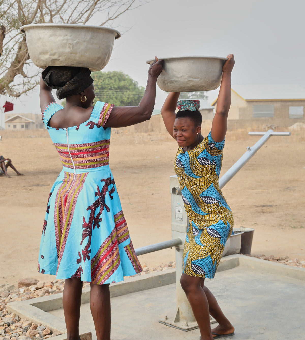 Two women in bright wax-print dresses lift metal basins onto their heads beside a new hand-pump water well in northern Ghana.