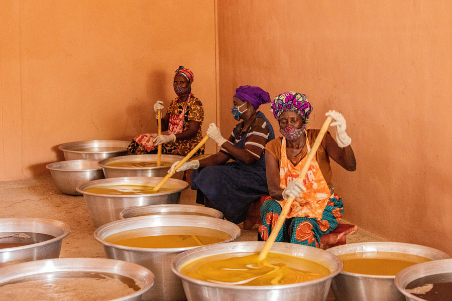 Three women in face masks and gloves sit among large metal basins of golden shea butter, stirring with long wooden paddles.