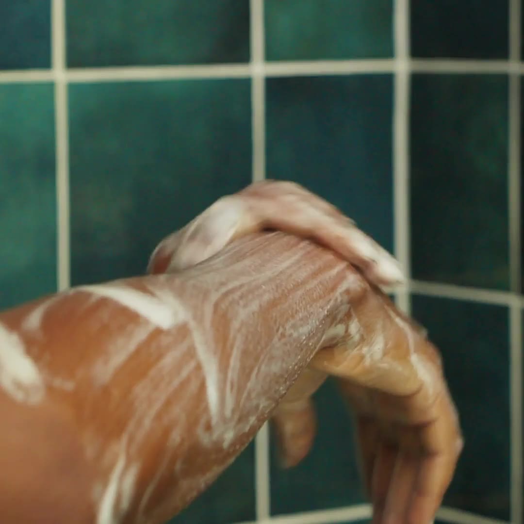 A soapy hand lathering body wash over the other forearm in front of green shower tile.