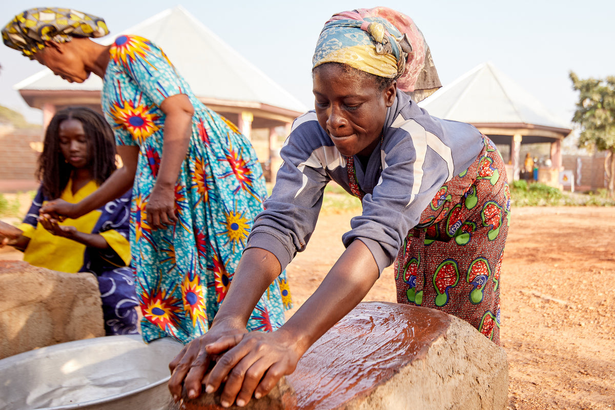 A woman in a grey jacket leans over a stone, kneading shea butter by hand while other women work behind her.