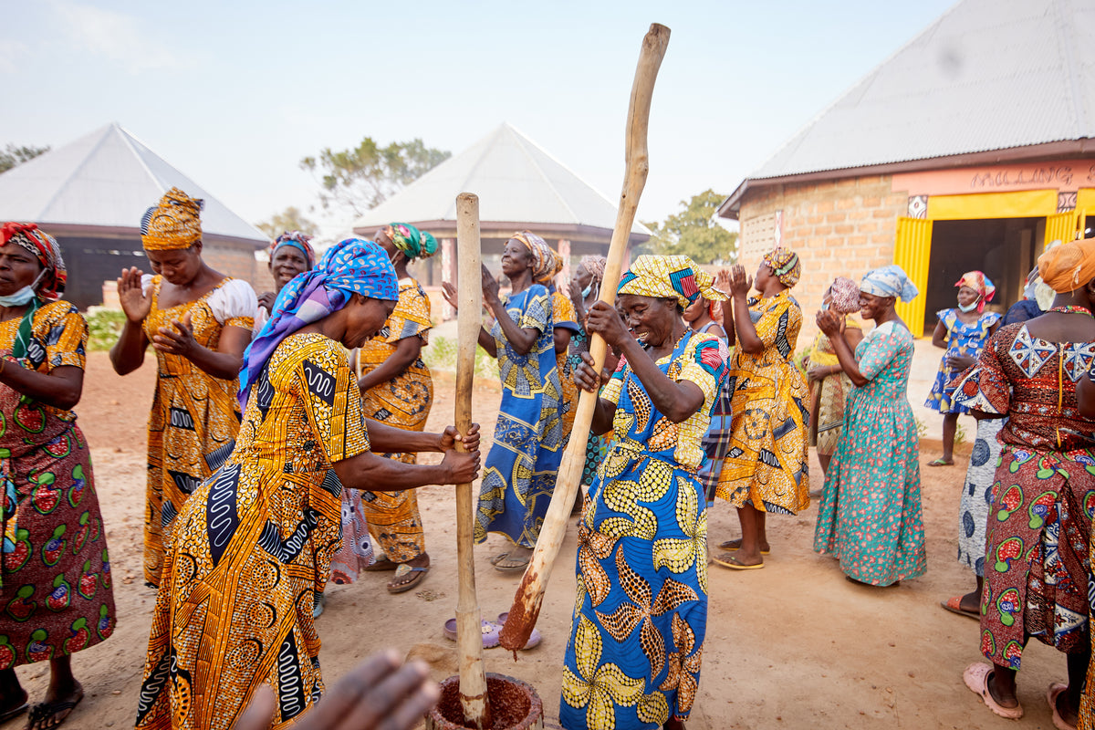 Women in bright wax-print dresses pound shea nuts with tall wooden pestles in a cooperative courtyard under a wide blue sky.
