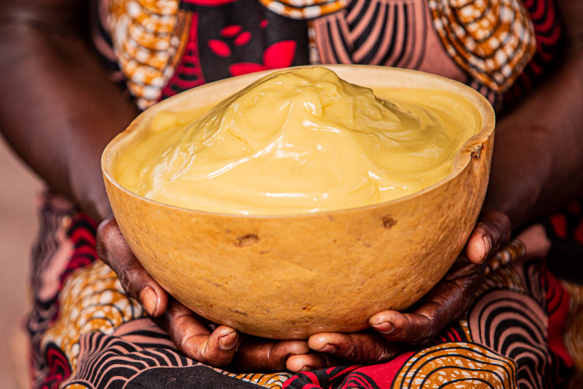 Hands holding a wooden bowl of golden shea butter in front of colorful African wax-print fabric.
